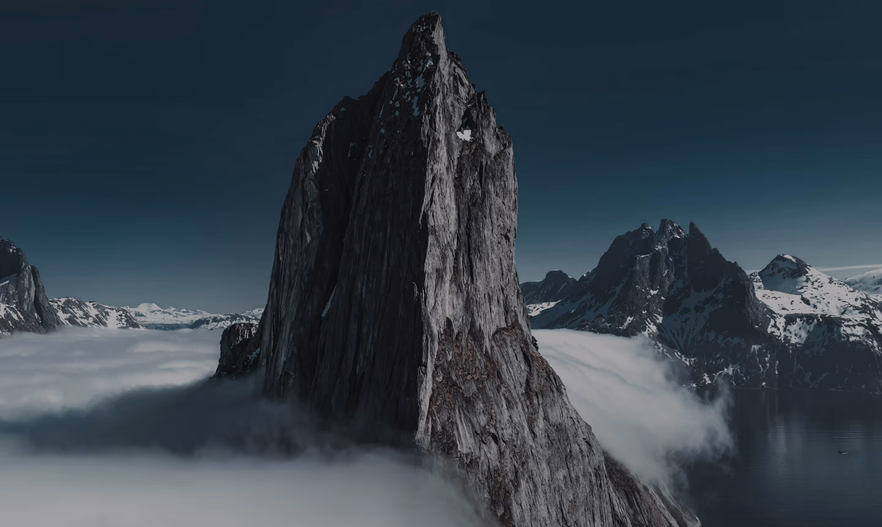 Sharp rocky mountain peak rising above low clouds with snow-covered mountains and calm water in the background under a dark sky.