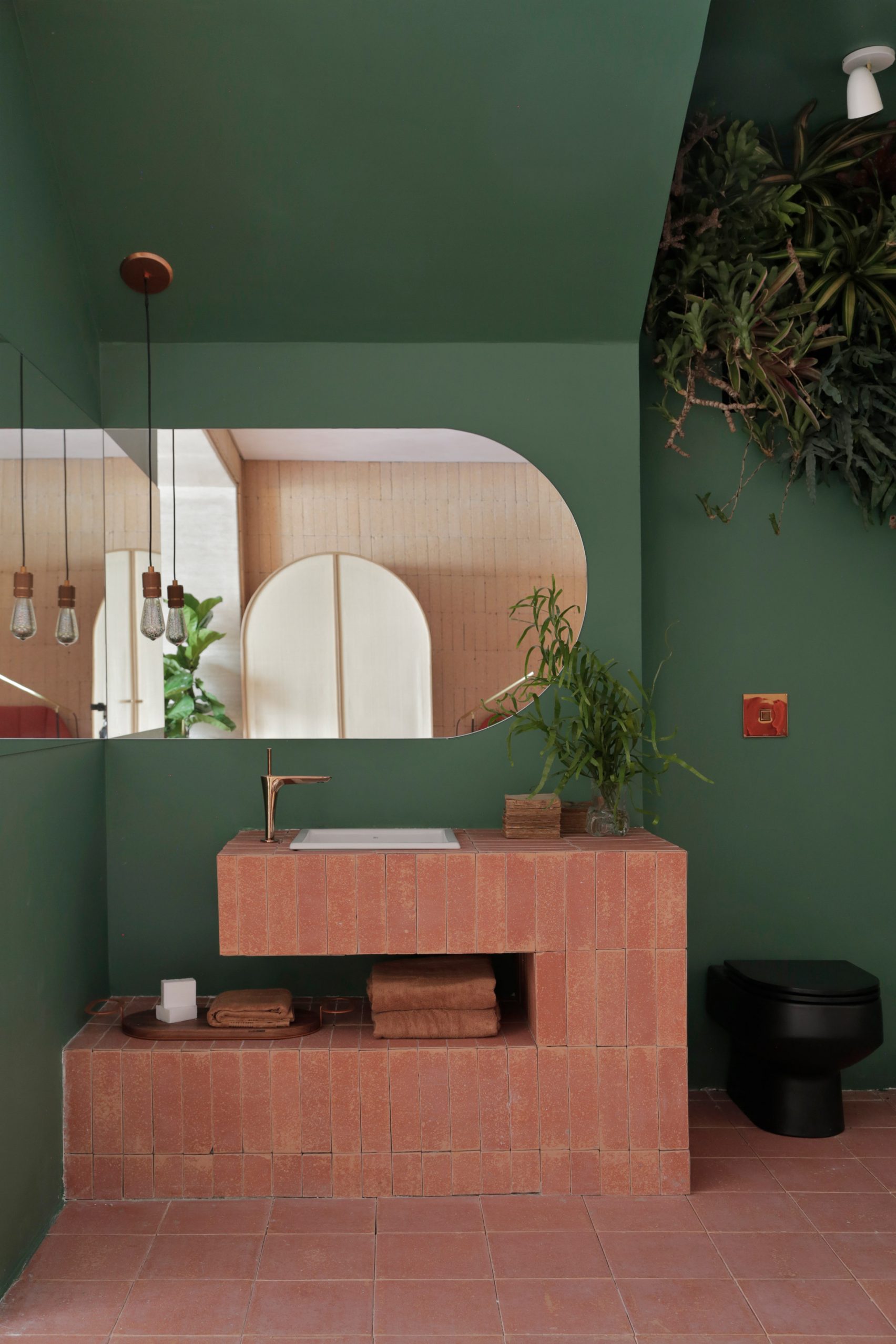 Bathroom with green walls, terracotta tiled sink, black toilet, plants, and a large rounded mirror.