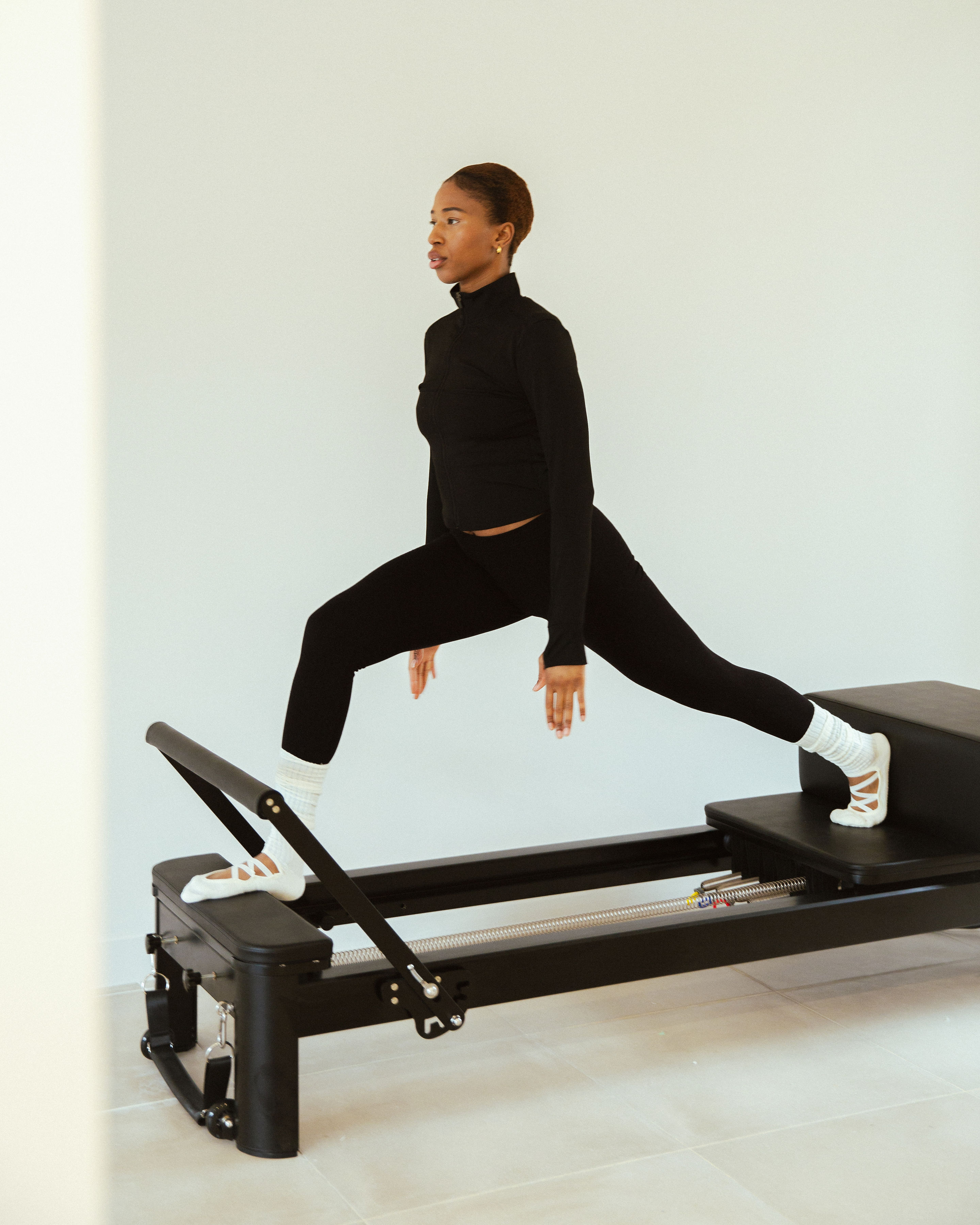 Woman in black activewear doing a Pilates lunge stretch on a reformer machine indoors.