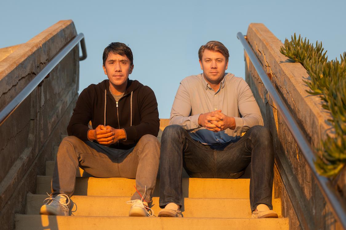 Two men sitting on outdoor concrete stairs with railings, hands clasped, looking at camera.
