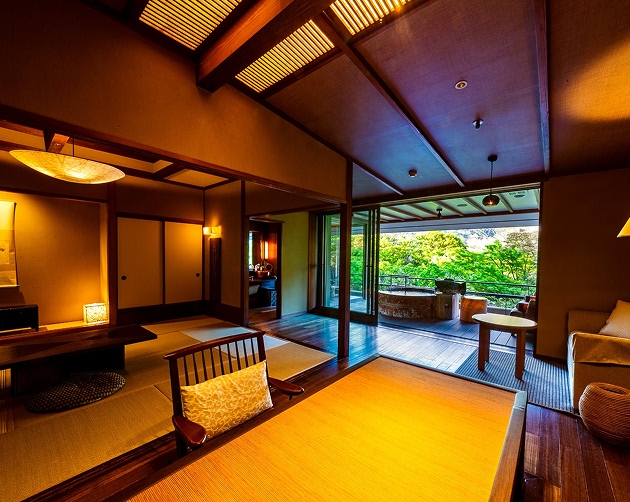 Warmly lit traditional Japanese room with tatami mats, wooden furniture, and sliding shoji doors, opening to a balcony with stone hot tub and green tree view.