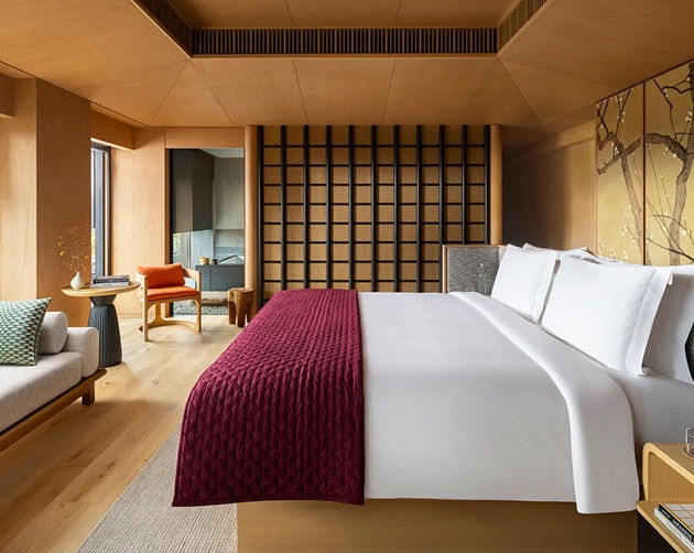 Modern hotel room with large bed covered in white linens and a deep red quilted throw, a wooden floor, beige walls, a decorative screen behind the bed, a small sitting area with an orange chair and a side table near a window, and a sofa with patterned pillows.