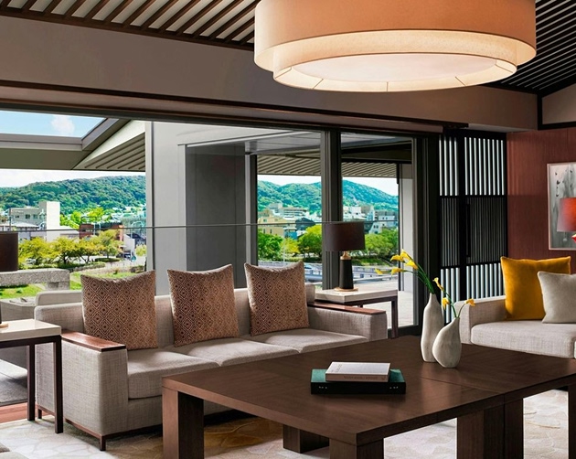 Modern living room with beige sofas adorned with patterned and solid cushions facing a dark wood coffee table holding two books and two white vases with yellow flowers, large windows showing green hills and buildings outside, and a large round ceiling light above.