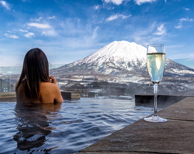 A person with long dark hair relaxing in an outdoor hot spring pool, facing a snow-capped mountain under a blue sky with scattered clouds, with a tall glass of champagne on the poolside.