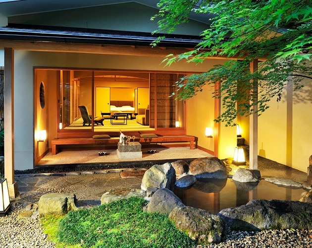 Evening view of a traditional Japanese-style room with sliding doors, warm interior lighting, and wooden deck overlooking a garden with a stone-lined pond and green maple tree branches.