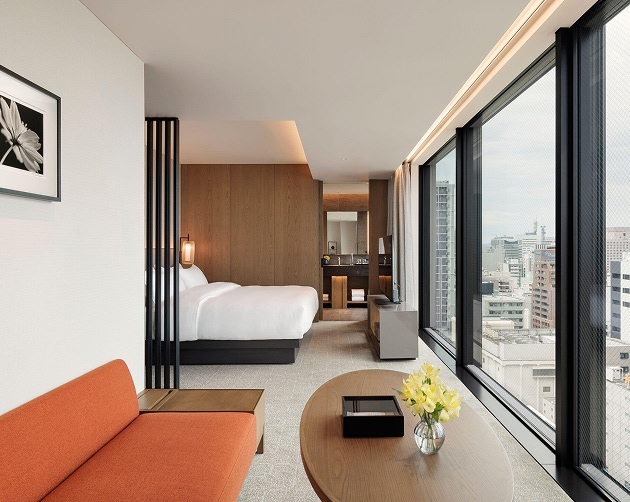 Modern hotel room with a large bed and wooden headboard, an orange sofa, a wooden oval coffee table with a glass vase of yellow flowers, and large windows offering a cityscape view.