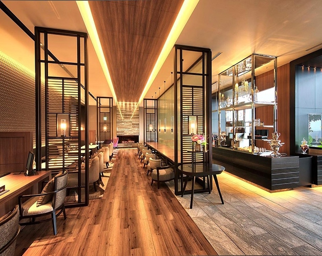 Modern, warmly lit lounge area with a long wooden floor pathway, comfortable armchairs arranged along the sides, black metal-framed glass partitions, and a sleek bar counter displaying glassware and floral decorations.