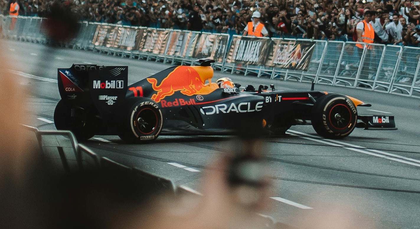 Red Bull Oracle Formula 1 race car making a sharp turn on a racetrack, with spectators behind barriers and people in orange safety vests watching.