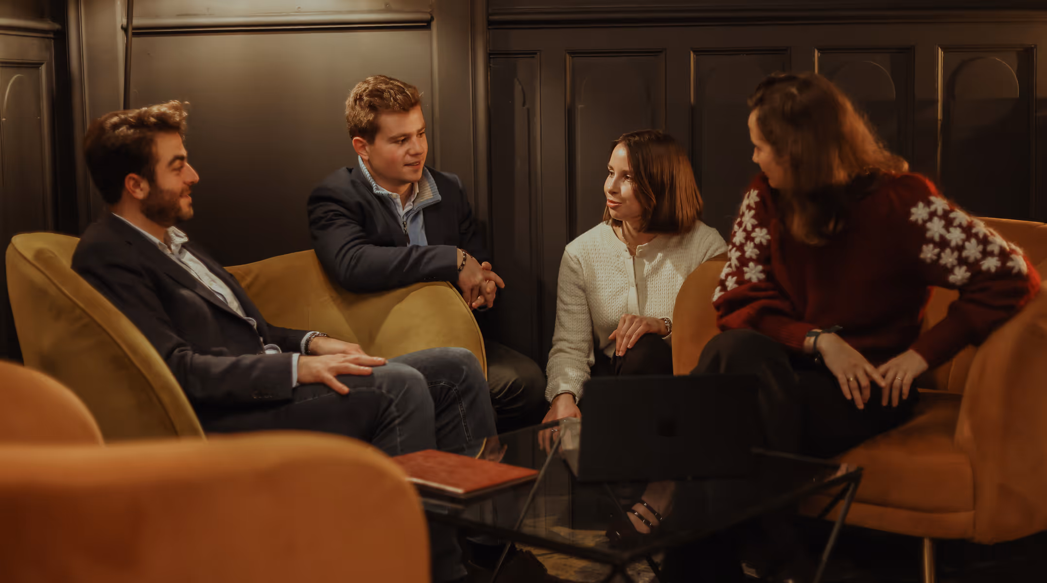 Quatre adultes discutent autour d'une table basse dans un salon avec des fauteuils confortables.