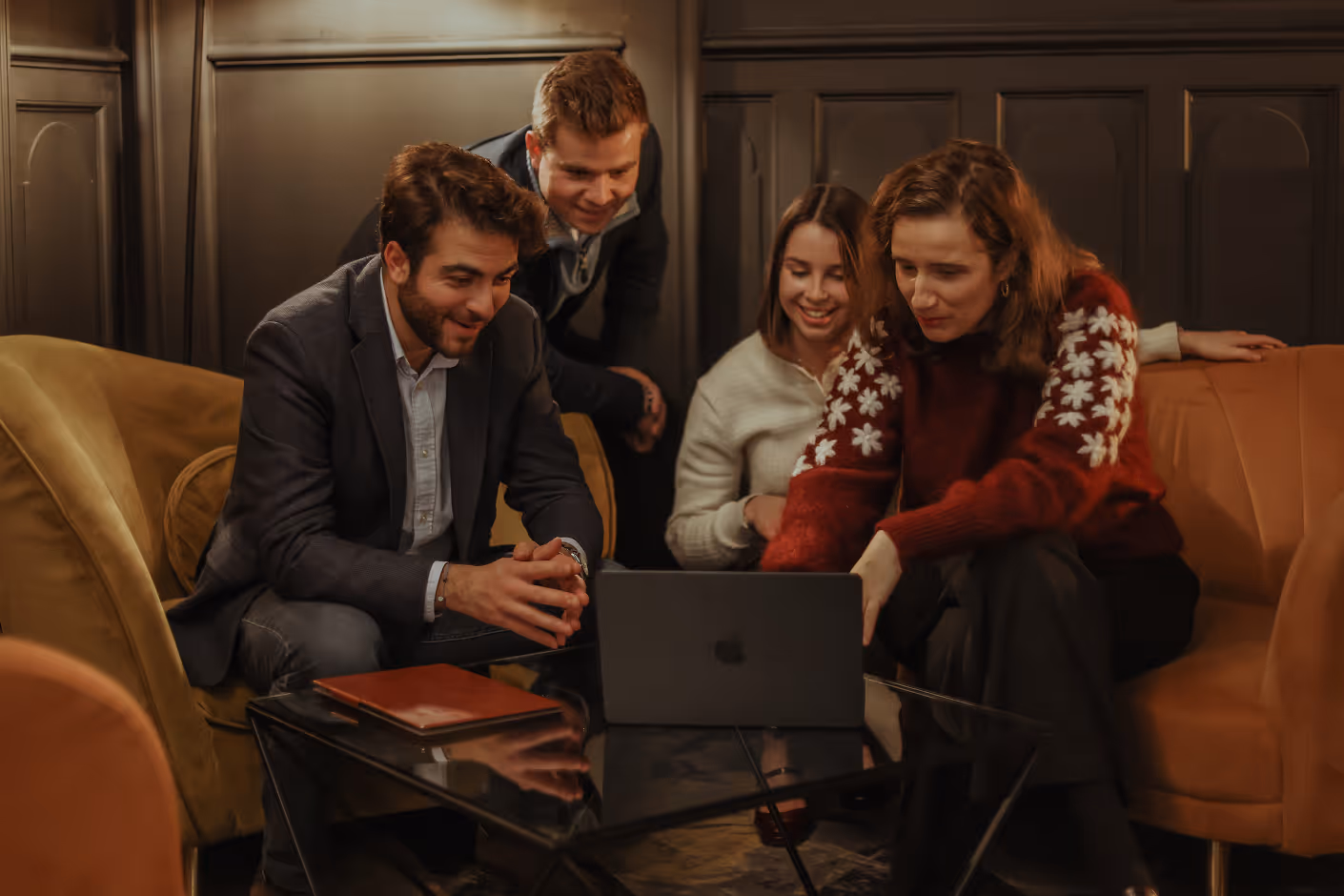 Quatre adultes souriants assis autour d'un ordinateur portable sur une table en verre dans un salon cosy.