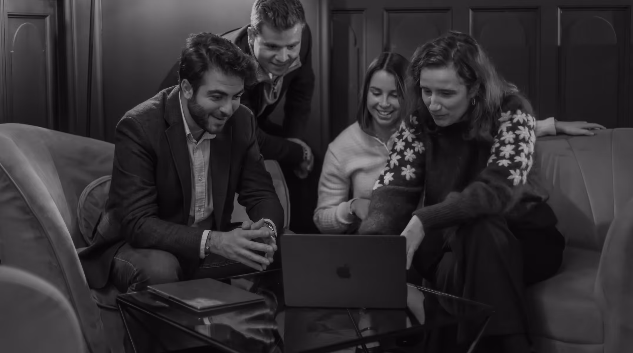Quatre personnes assises et debout autour d'une table avec un ordinateur portable, regardant l'écran et souriant.