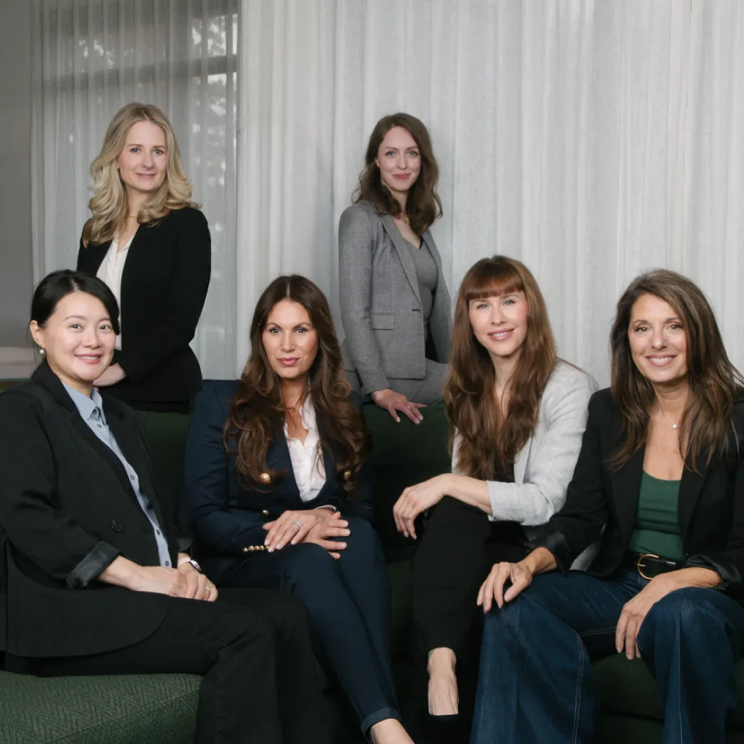 Six professionally dressed women pose together for a group portrait in an office setting.