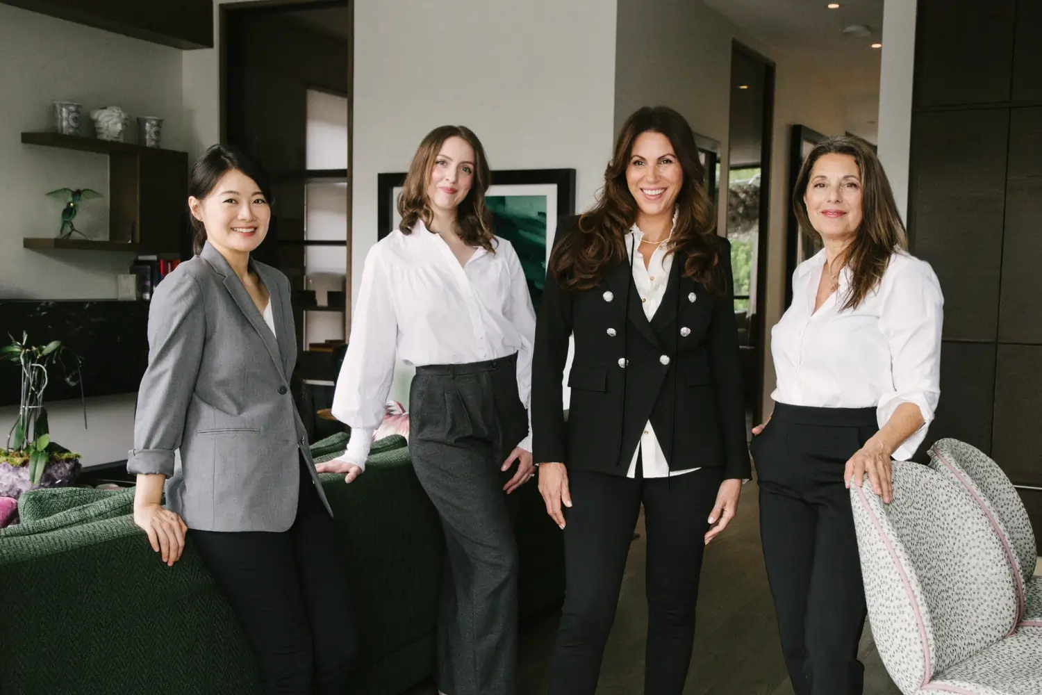 Four professionally dressed women standing together and smiling in a modern office.