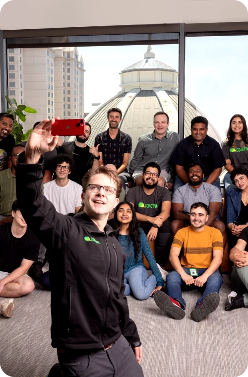 A diverse group of Coactive team members takes a selfie, with a man at the center, all displaying happy expressions and enjoying the moment.