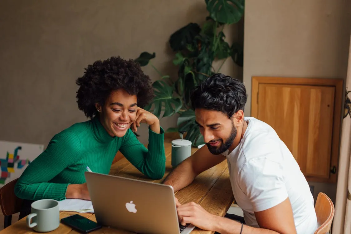 Young man and woman sitting at a wooden table looking at a laptop together, smiling and engaging in a cozy indoor setting.