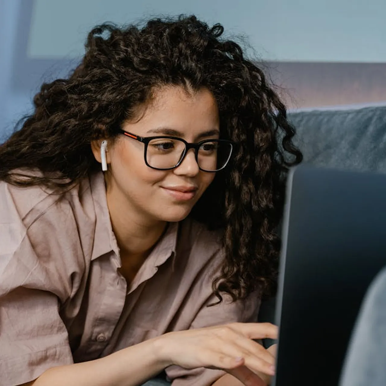 Young woman with curly hair and glasses using a laptop while wearing a wireless earbud.