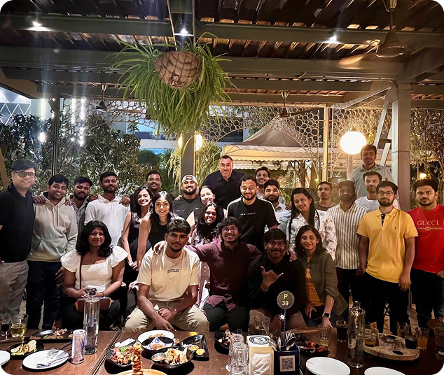 Group of young adults posing together under a canopy at an outdoor dining area with plates of food on the table in front.