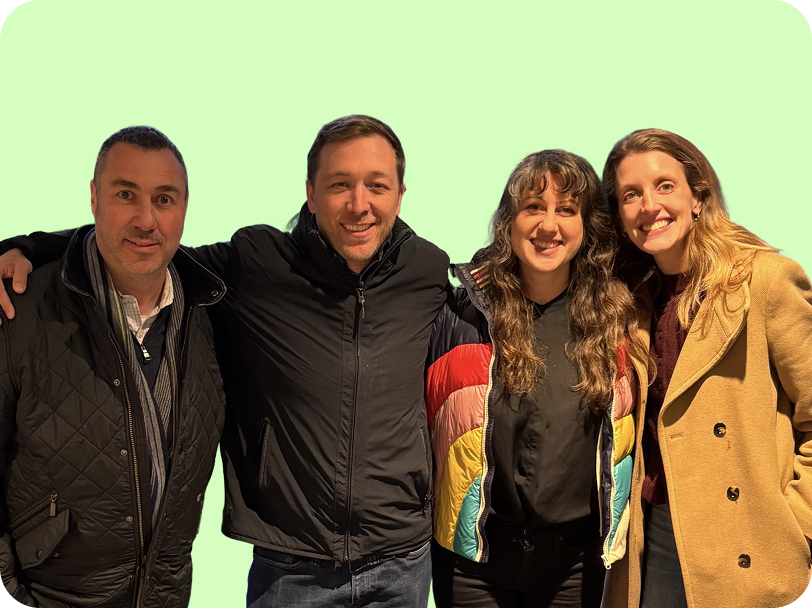 Four people standing close together smiling, wearing jackets and coats against a light green background.
