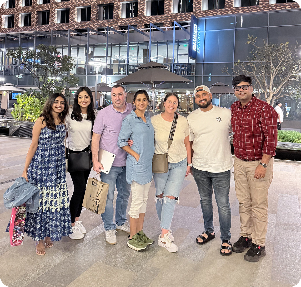 Seven people standing together outdoors at night in casual clothes, smiling at the camera with a modern building and patio umbrellas in the background.