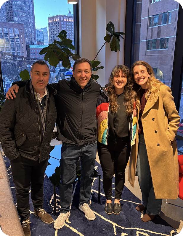 Group of two men and two women standing indoors in front of large windows with city buildings outside, smiling and posing with arms around each other.
