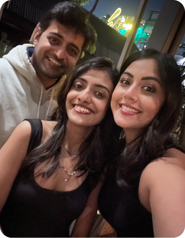 Three young adults smiling closely together in a nighttime indoor setting, two women in black tops and one man in a light-colored hoodie.