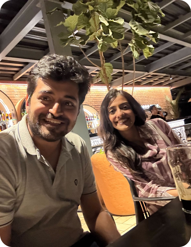 Man and woman smiling and sitting at a table in a warmly lit restaurant with a tall plant and bottles on shelves in the background.