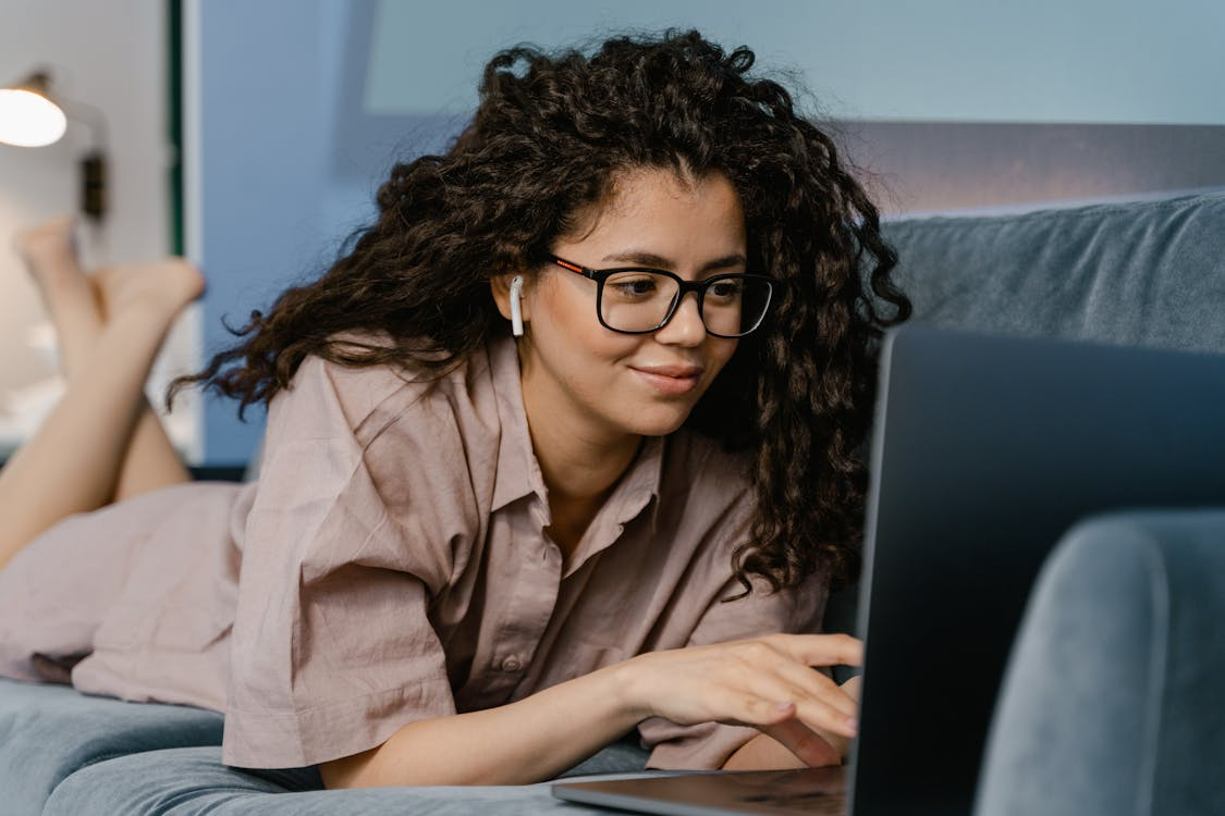 Young woman with curly hair and glasses lying on a couch using a laptop with wireless earphones.
