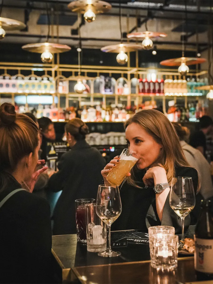 Zwei Frauen sitzen an einem Tisch in einer Bar, eine trinkt Bier, im Hintergrund sind Regale mit Flaschen und hängende Lampen.