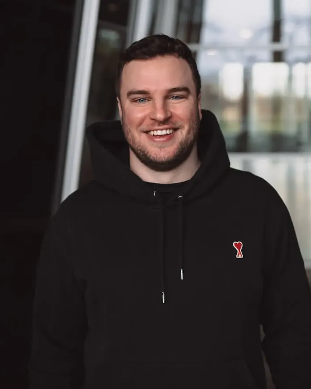 Lächelnder Mann mit kurzen dunklen Haaren und Bart trägt schwarzen Kapuzenpullover mit rotem Herzlogo.