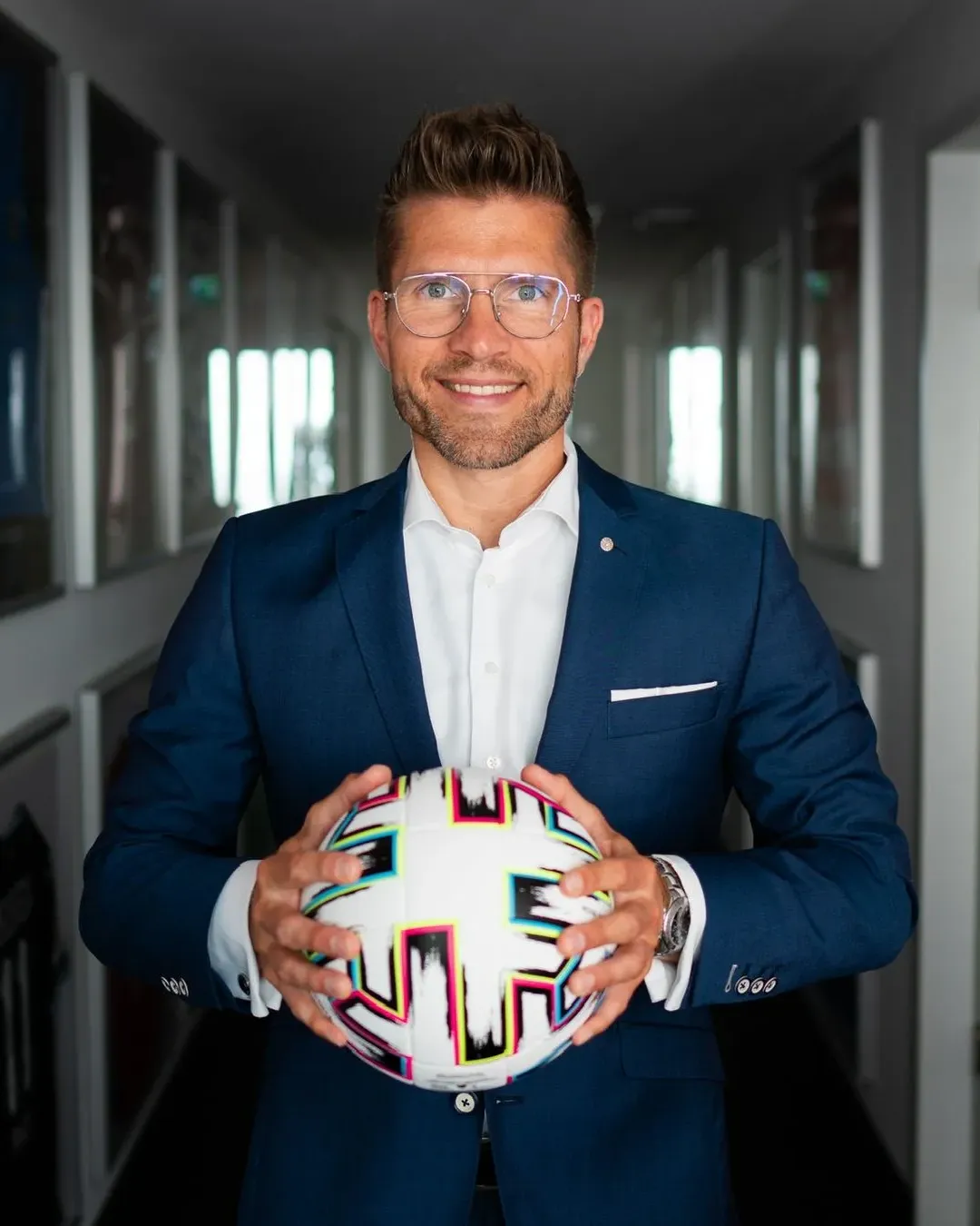 Mann mit Brille und blauem Anzug lächelt und hält einen bunten Fußball mit beiden Händen.