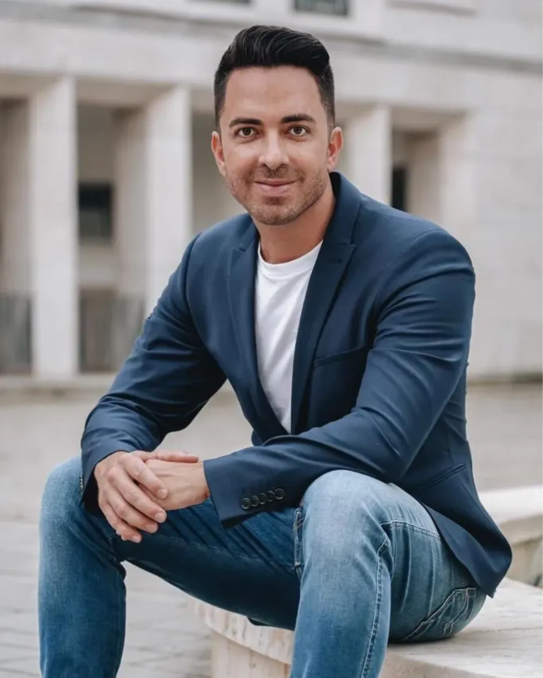 Junger Mann mit kurzem, dunklem Haar trägt blaues Sakko und Jeans, sitzt draußen auf einer Steinbank und lächelt freundlich.