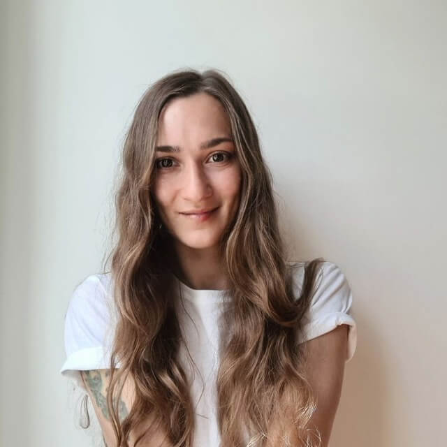 Жінка у білій футболці сидить біля світлої стіни та дивиться в камеру.
