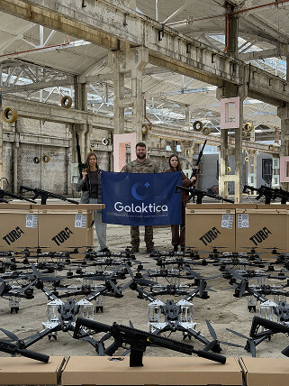 Team representatives holding a Galaktica flag in front of donated FPV drones and shotguns