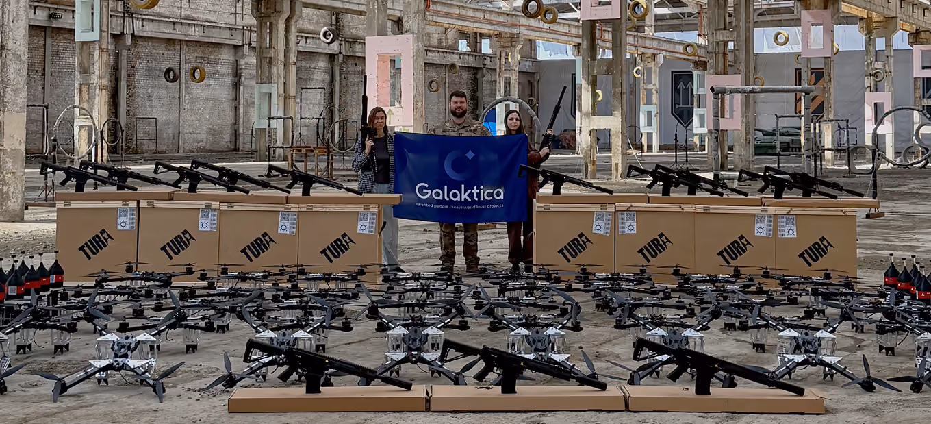 Team representatives holding a Galaktica flag among donated FPV drones, shotguns, and equipment