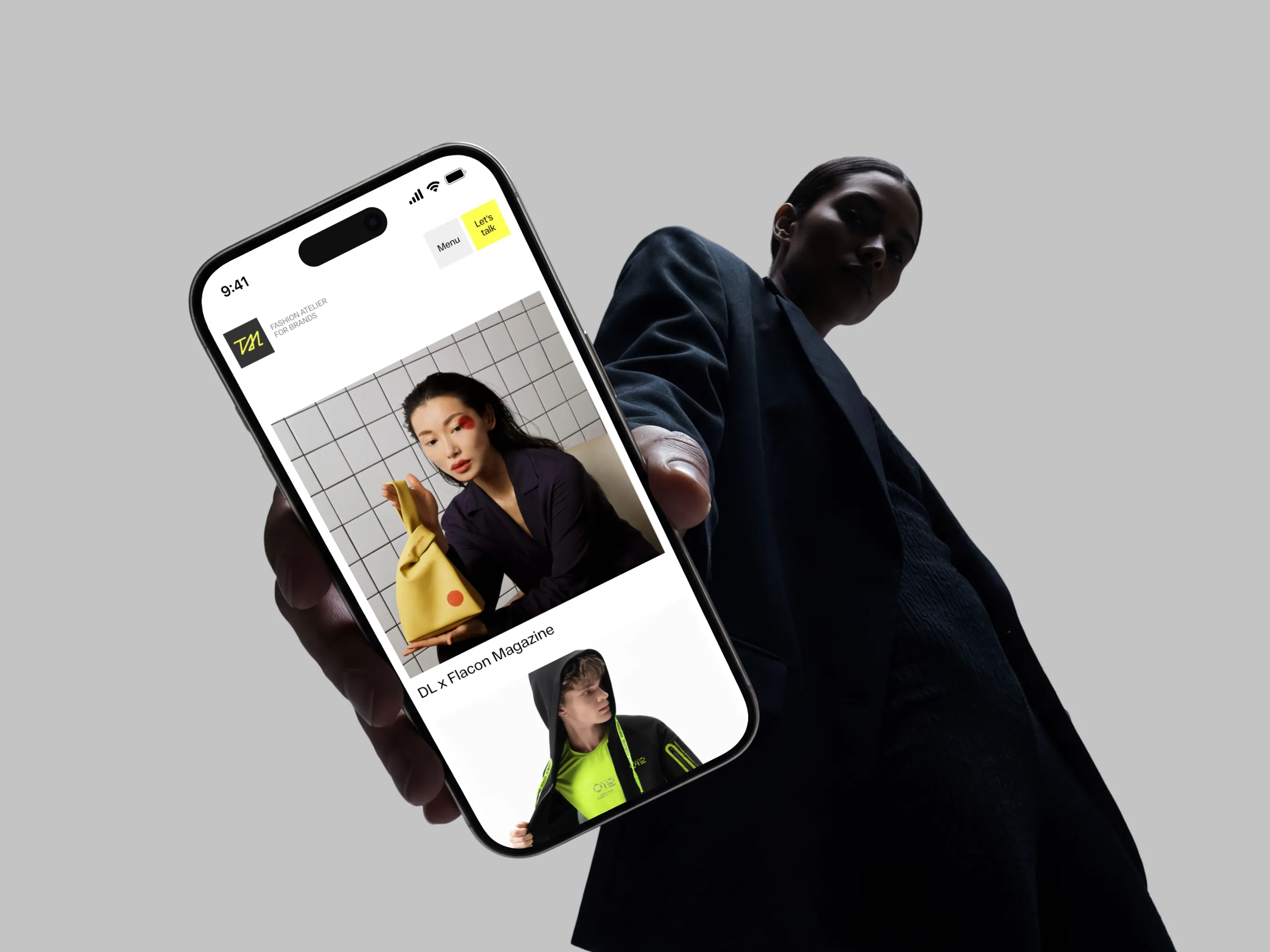 Person holding phone showing fashion website with models and a yellow handbag against gray background.