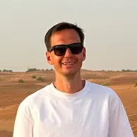 Smiling man wearing sunglasses and white shirt standing in a desert landscape with a clear sky.