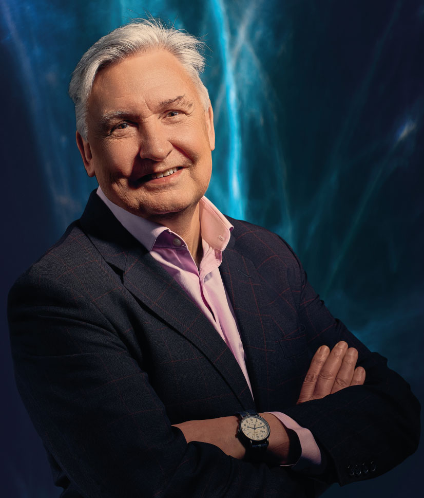 Older man with gray hair wearing a dark suit jacket and light pink shirt, smiling with arms crossed against a dark background with blue light patterns.