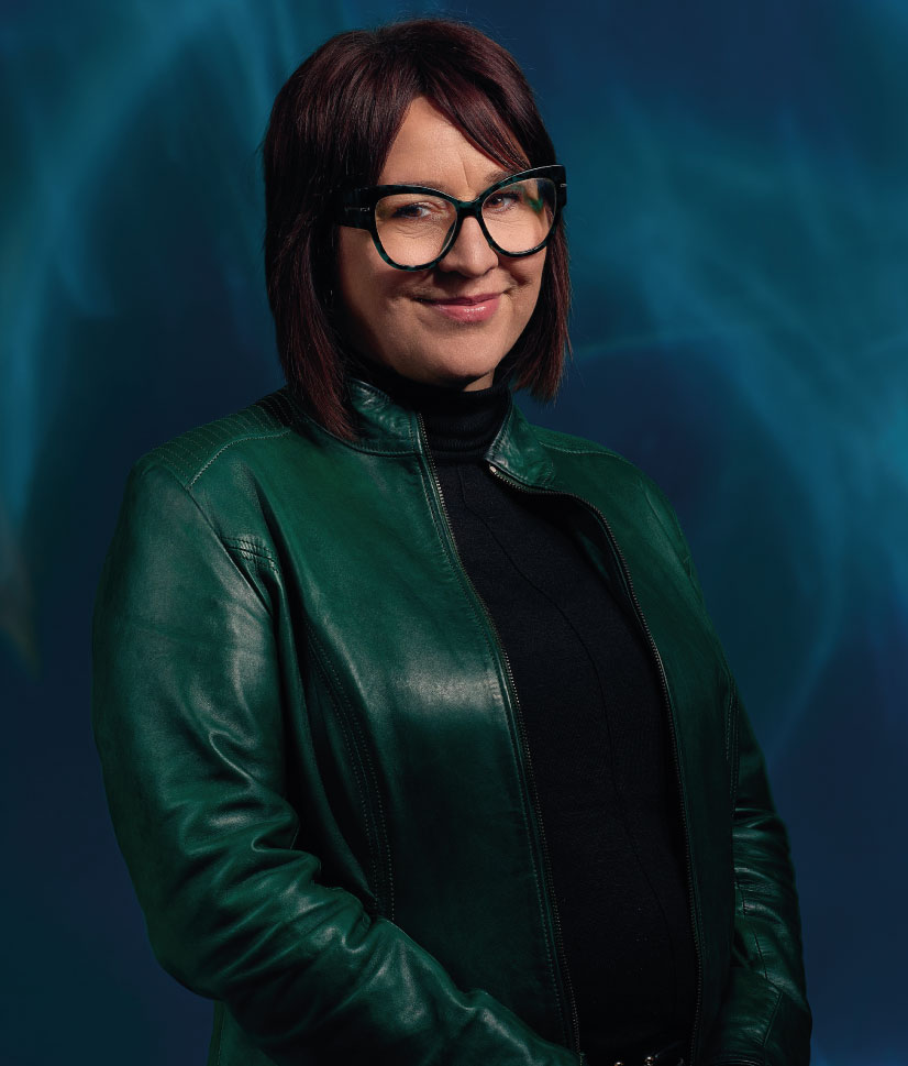 Woman with medium-length dark hair and large black-rimmed glasses, wearing a green leather jacket over a black top, posing against a dark blue background.