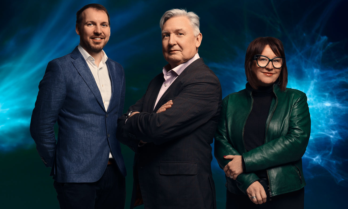 Three professionally dressed adults posing against a dark blue abstract background: a smiling man in a blue blazer and white shirt on the left, an older man with gray hair in a dark suit jacket and light pink shirt with arms crossed in the center, and a woman with short dark hair, glasses, and a green leather jacket over a black top on the right.