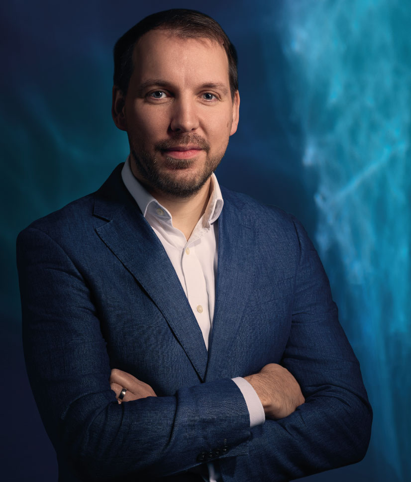 Man with short dark hair and beard wearing a blue blazer and white shirt, standing with arms crossed against a dark blue and turquoise blurred background.