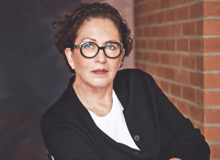 Marianne Leber, collage artist, portrait photograph with glasses seated against a brick wall.
