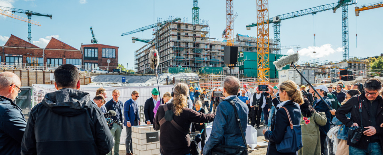 Gruppe von Menschen bei einer Grundsteinlegung auf einer Baustelle mit Kränen und Gebäuden im Hintergrund.