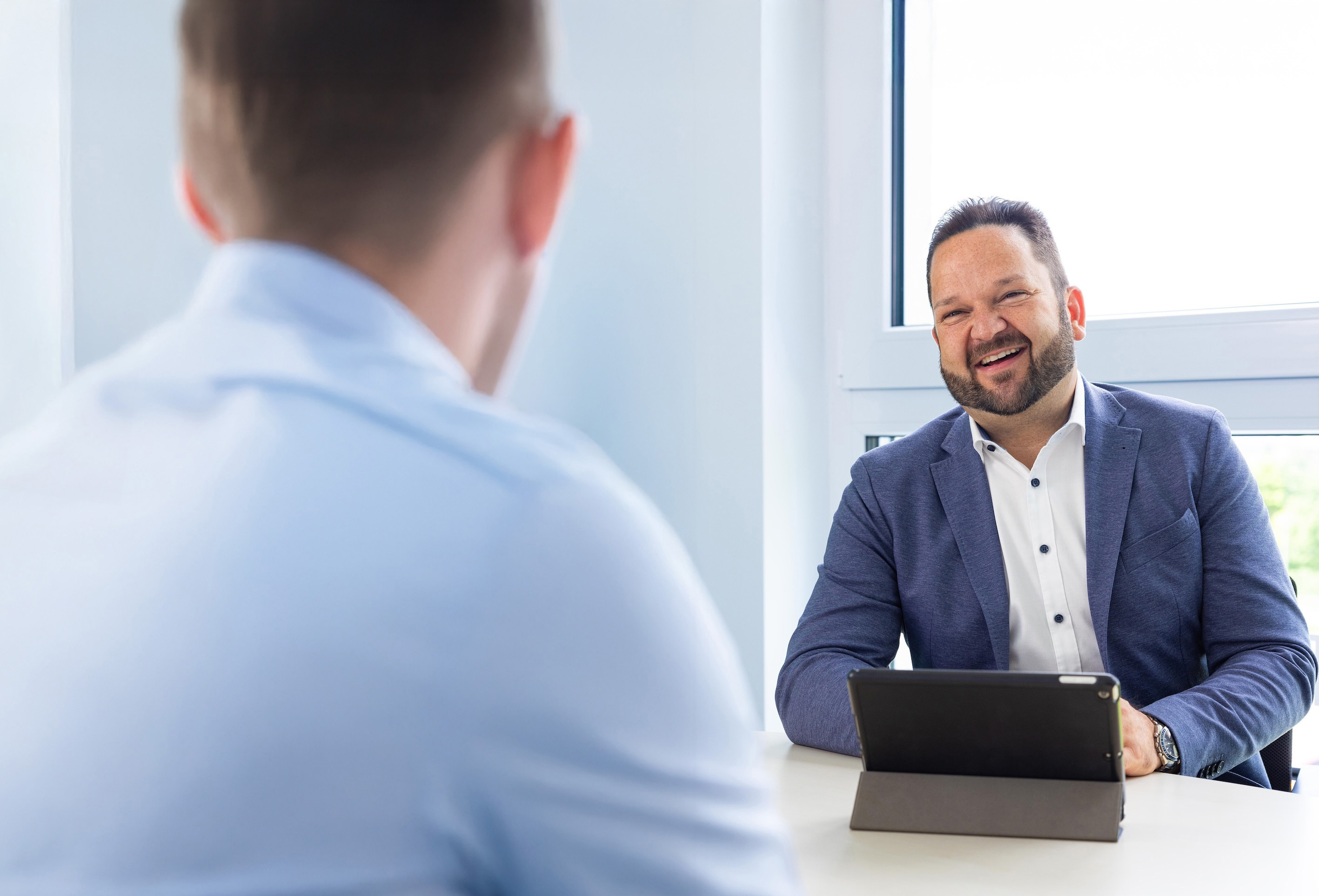 Geschäftsmann in blauem Anzug lächelt während eines Gesprächs mit einer anderen Person in einem hellen Büro.
