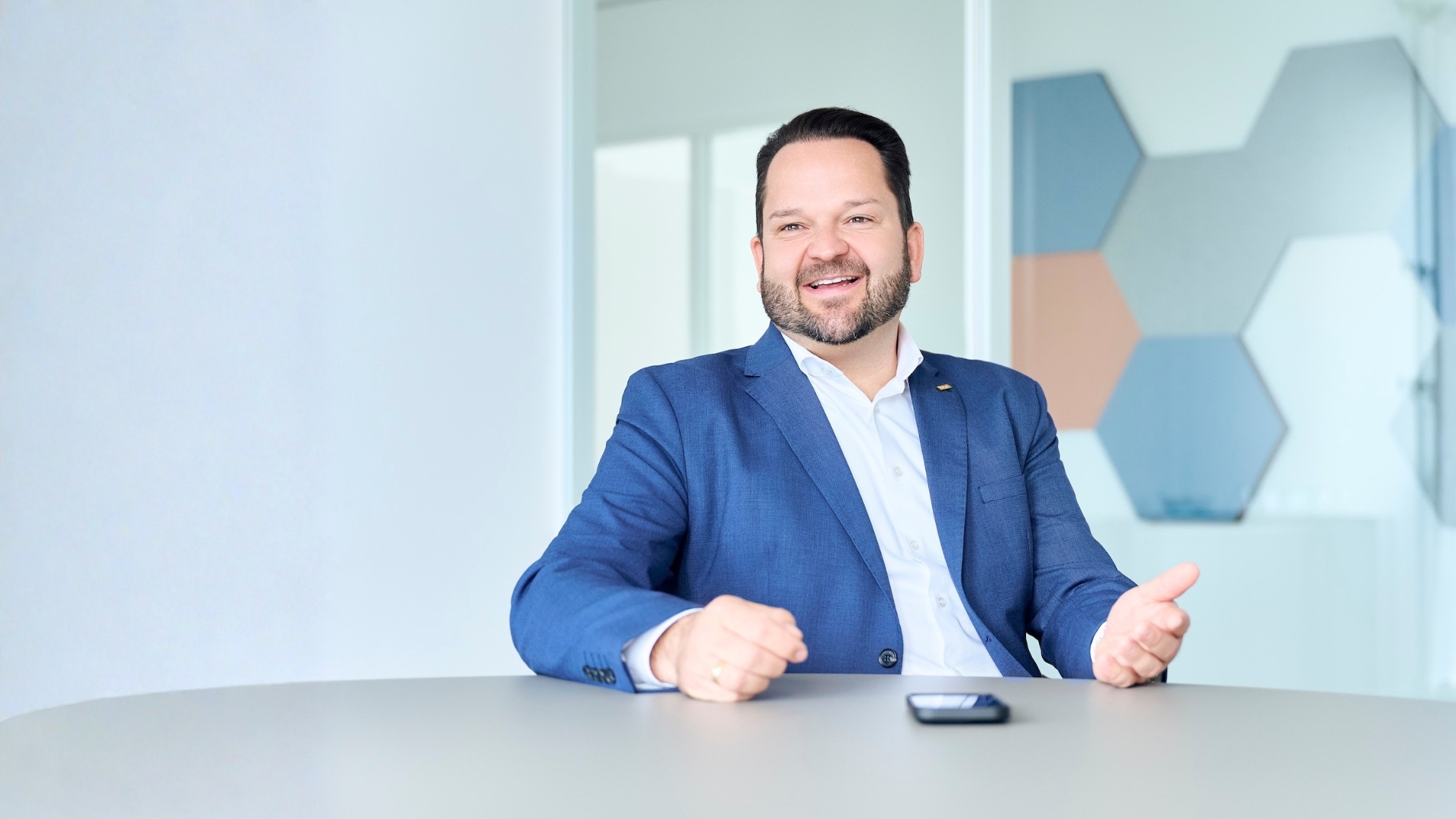 Mann mit dunkelblauem Anzug und weißem Hemd sitzt an einem Tisch und spricht lachend.