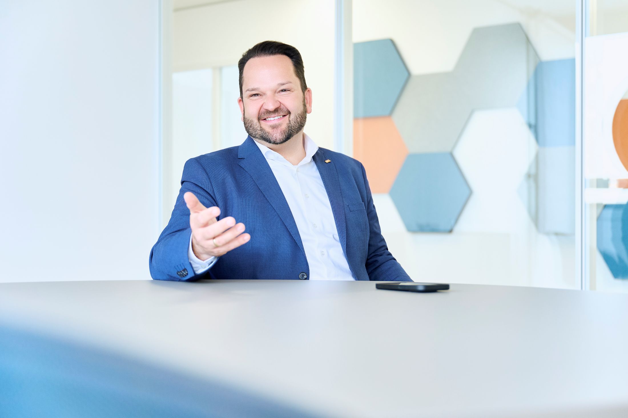 Ein lächelnder Mann in blauem Anzug sitzt an einem Tisch mit ausgestreckter Hand und einem Smartphone darauf.