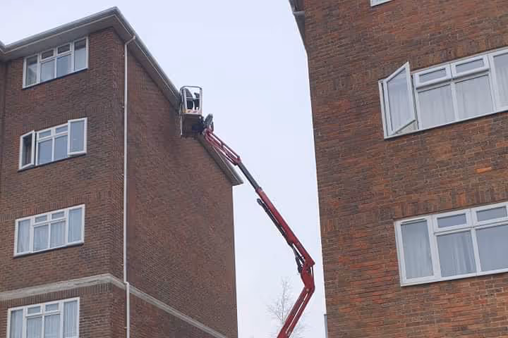 A person on a crane fixes the roof of a tall brick building.