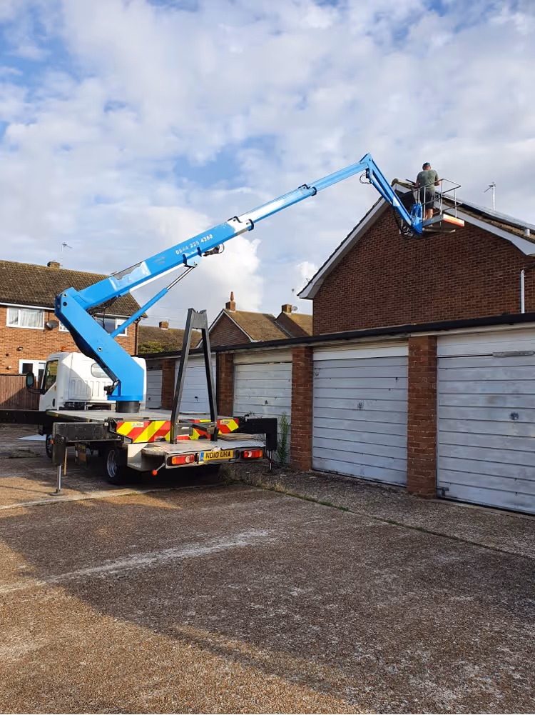 A worker on a blue cherry picker repairs the roof of a brick garage building.