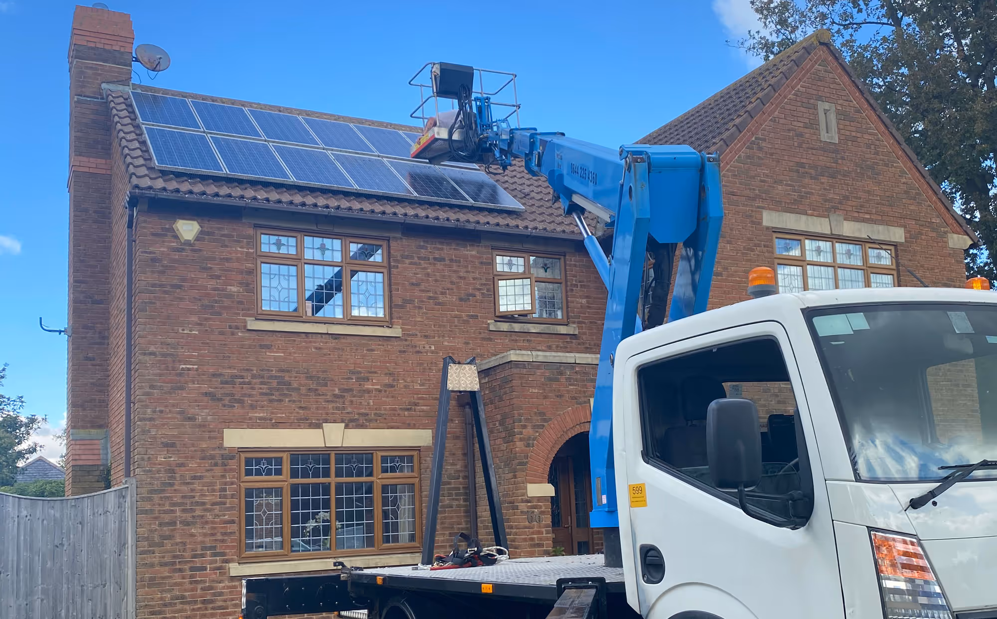 A worker in a lift installs solar panels on the roof of a brick house.