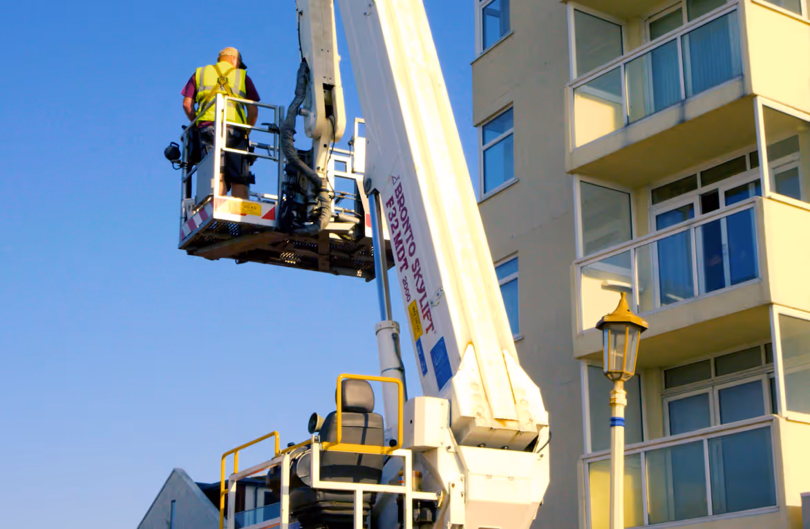 A worker in a lift is fixing a light next to a tall building.
