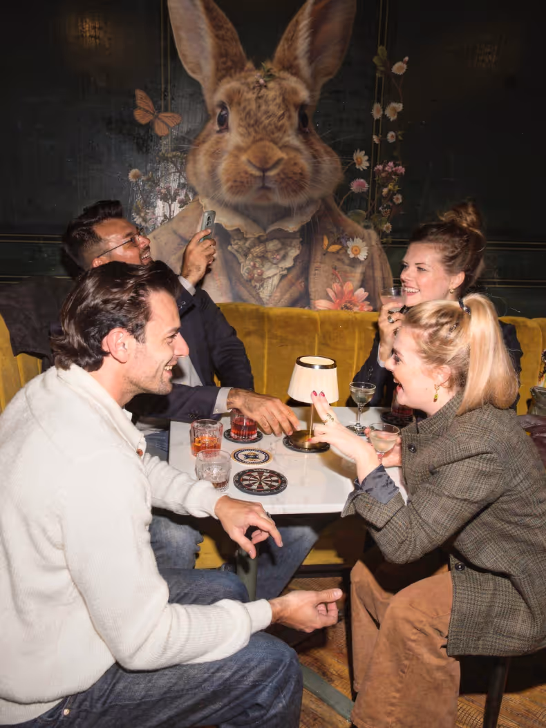 Four people sitting around a table in a cozy bar, smiling and holding drinks, with a large artistic mural of a rabbit wearing vintage clothing on the wall behind them.